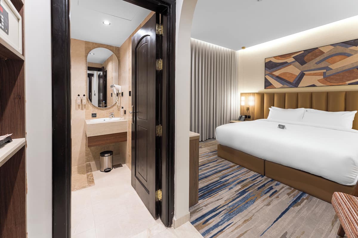 A hotel room with a bed, a bathroom with a mirror, sink, and toilet, a trash bin, and a wall-mounted artwork.