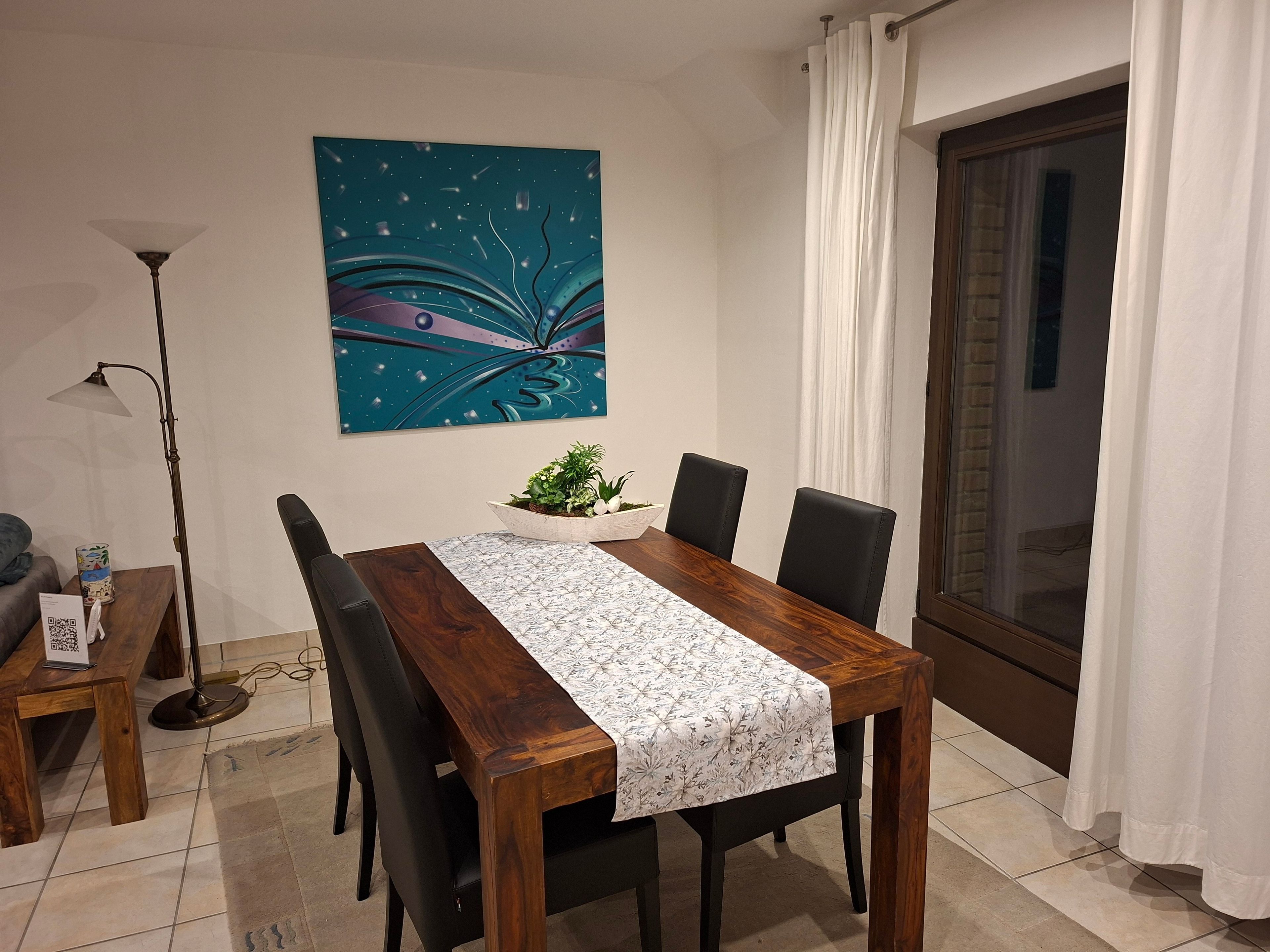 A dining area with a wooden table, black chairs, a decorative wall piece, a floor lamp, and a view through a window.