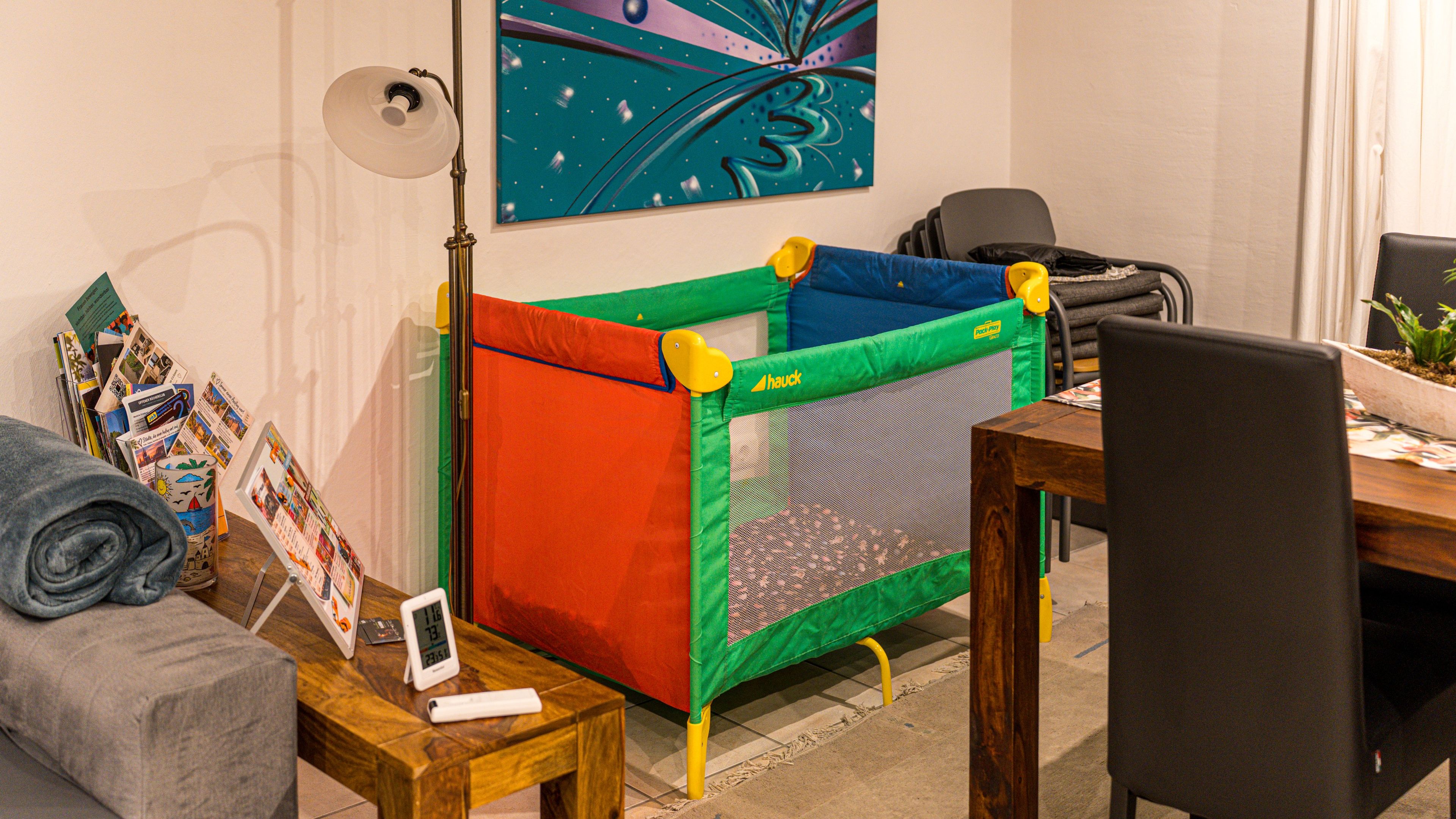 Ein Zimmer mit einem Spielbereich, einem Schreibtisch mit Lampe, einem Stuhl, einer Couch und einem Wandbild.