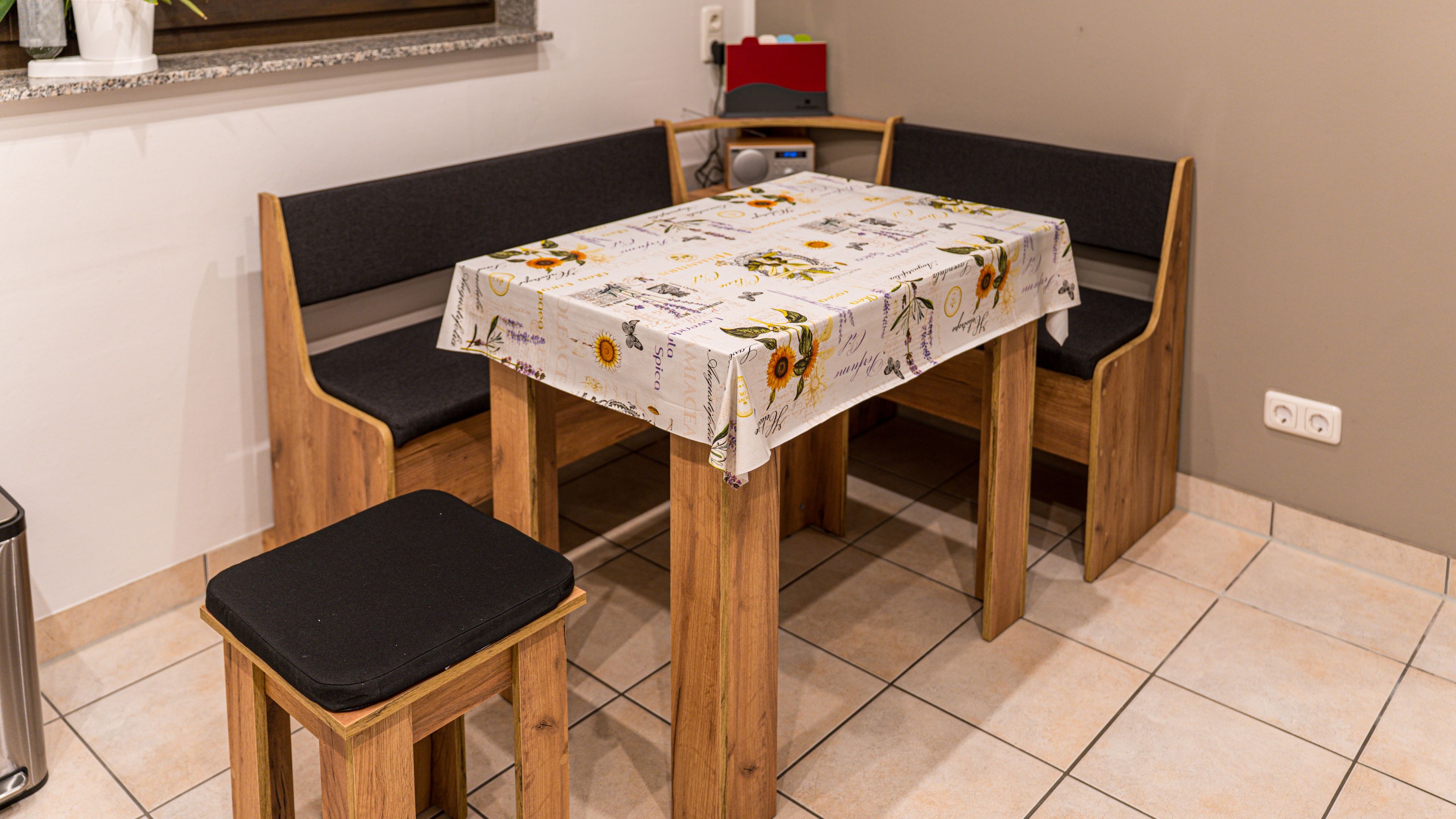 A small dining area with a table covered by a floral tablecloth, two wooden chairs with black cushions, and a bench.