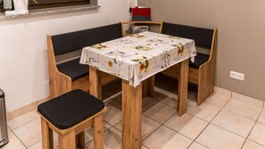 A small dining area with a table covered by a floral tablecloth, two wooden chairs with black cushions, and a bench.