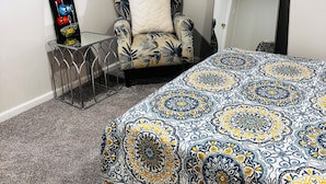 A room with a patterned bedspread, a chair with a floral design, a metal side table, and a wall-mounted decorative piece. - Cozy room in delightful Wilmington home, close to all local attractions (Wilmington)
