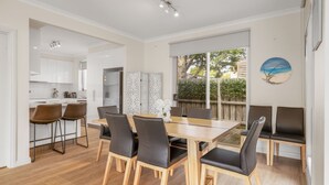 A dining area with a wooden table and chairs, a kitchen in the background, and a view of the outdoors through a large window. - Fair Haven in Cape Woolamai (Cape Woolamai)