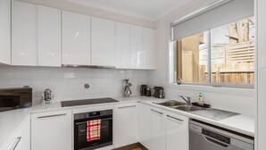 A modern kitchen with white cabinets, a built-in oven, a dishwasher, and a window with a view of a wooden fence. - Fair Haven in Cape Woolamai (Cape Woolamai)