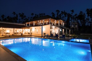 Ein Swimmingpool mit Liegestühlen und einer überdachten Zone mit Beleuchtung, umgeben von Palmen.