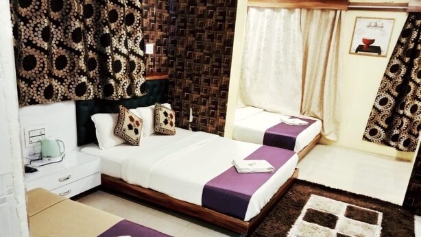 A bedroom with a double bed, a bedside table, a small desk, a chair, a patterned wall, and a window with curtains. - Hotel Royal Inn By Glitz (Mangaluru)