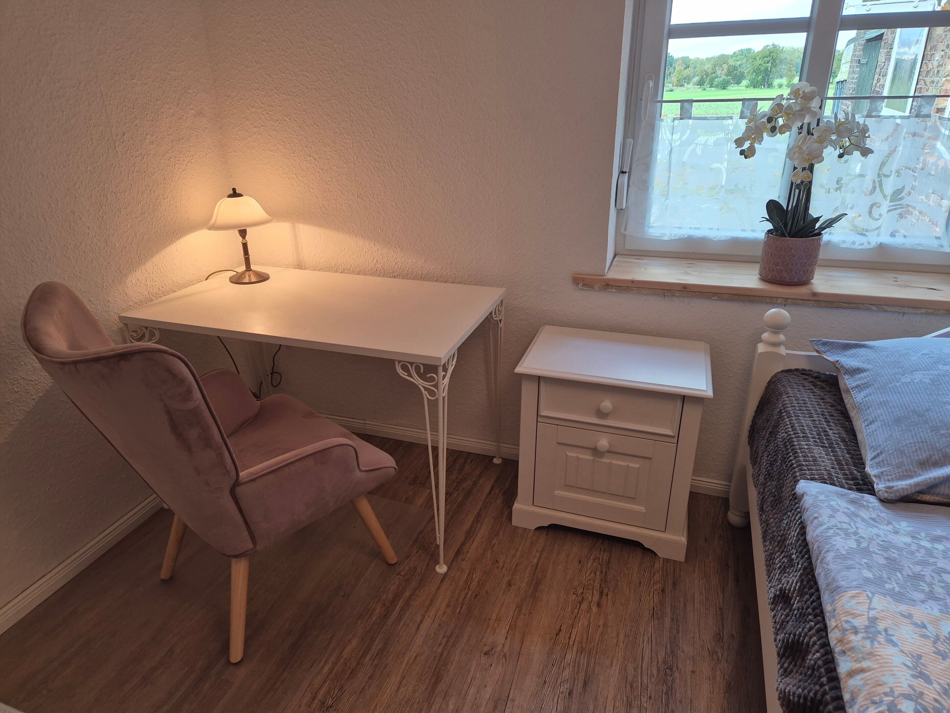 Un dormitorio con suelo de madera, un escritorio blanco con lámpara, una silla rosa, una mesita de noche con cajón, una cama con manta estampada y una ventana con vistas a zonas verdes.