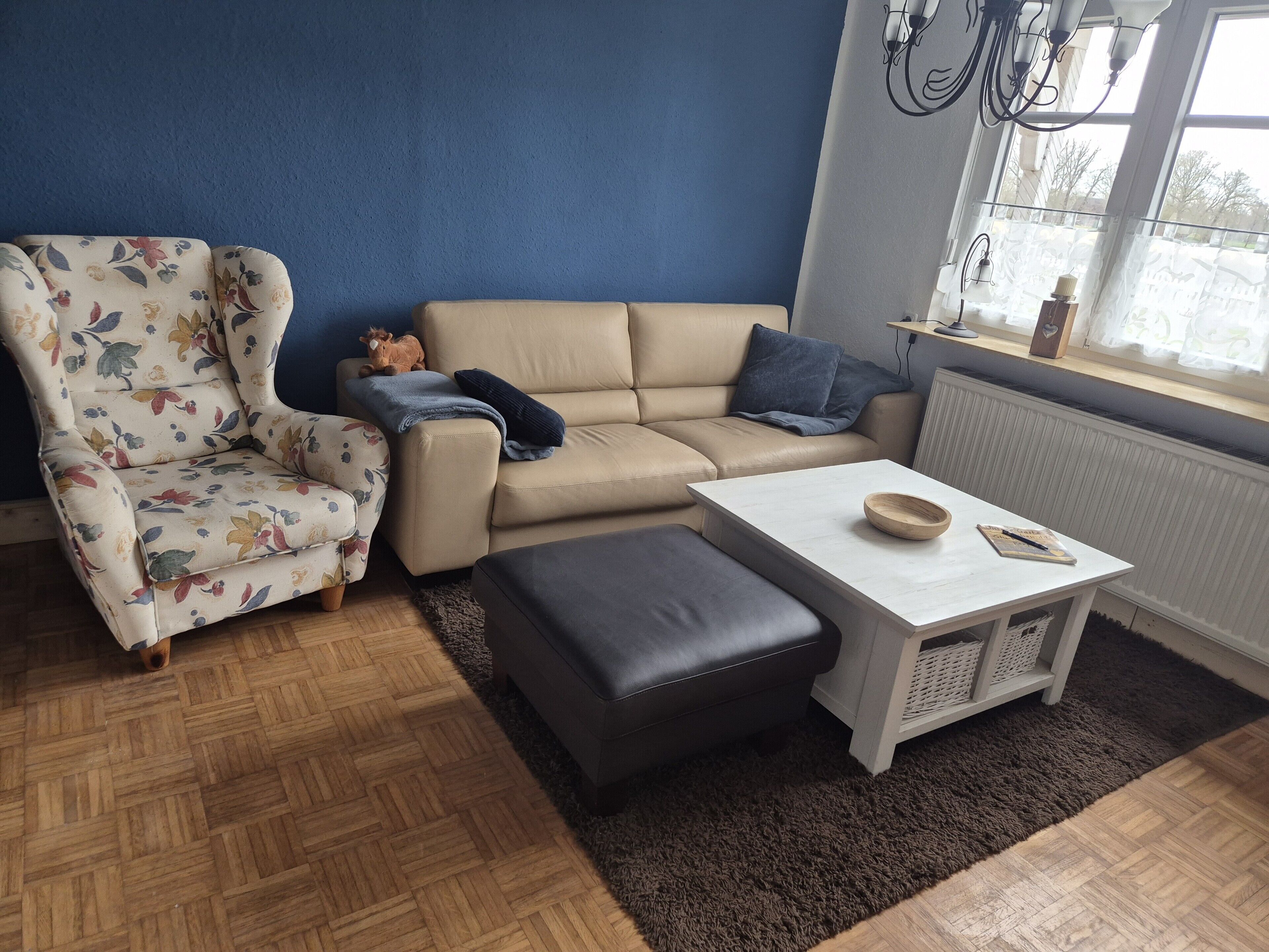 Una sala de estar con un sillón estampado, un sofá beige, un reposapiés oscuro, una mesa de centro blanca y un ventanal con vistas.