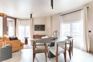 A modern living room with a round dining table, comfortable seating, a flat-screen TV, and large windows with curtains. - Appartement de luxe vue sur la Madeleine (Paris)