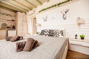A bedroom with a bed, pillows, a bedside table, a nightstand, a wall-mounted lamp, and a shelf with decorative items. - Opus Studio by Mykonian Hospitality (Mykonos)