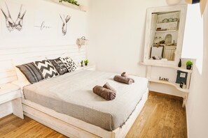A bedroom with a bed, pillows, a nightstand, a bedside table, a mirror, a shelf with a plant, and a wall with artwork. - Opus Studio by Mykonian Hospitality (Mykonos)