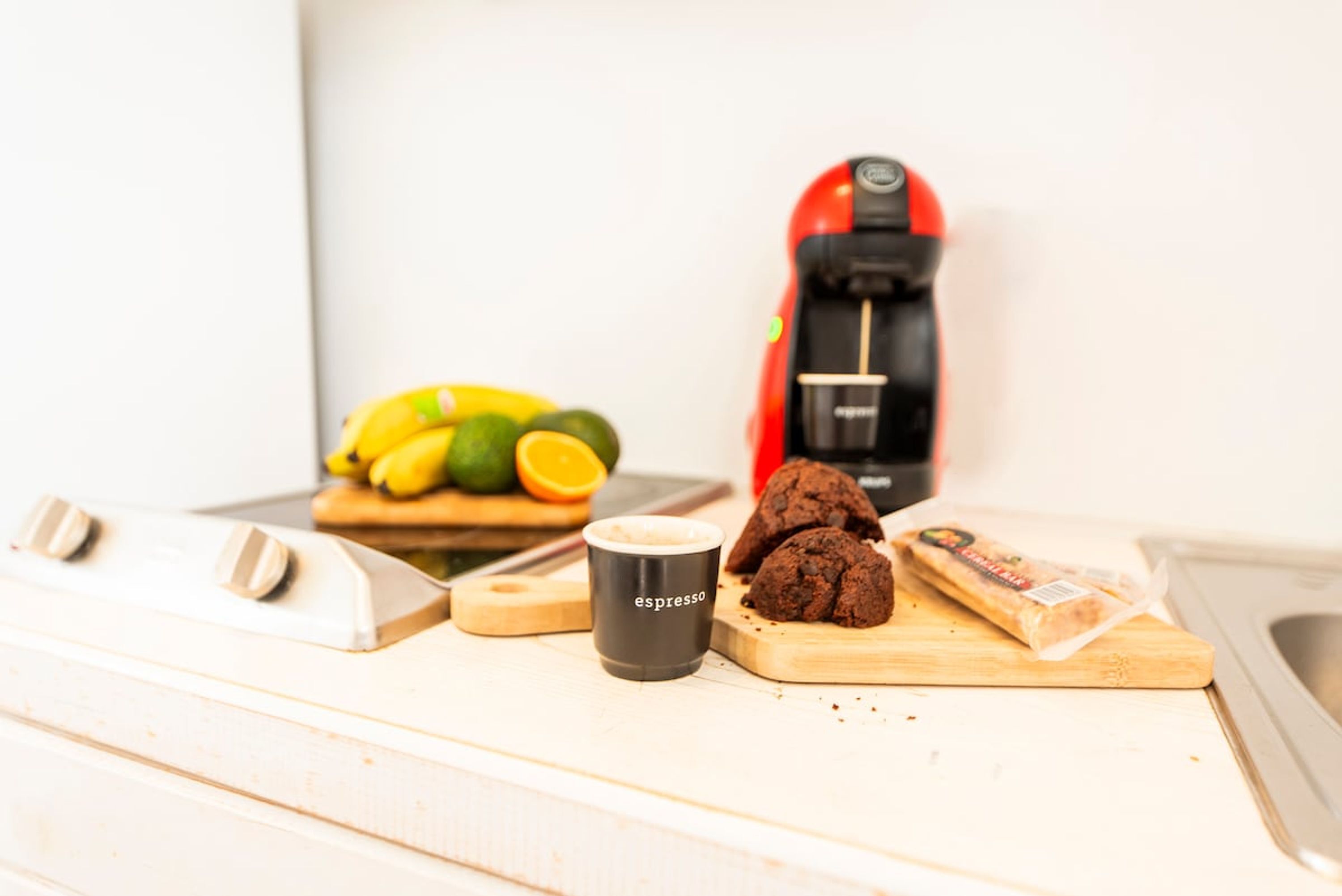 A kitchen counter with a coffee maker, a cup labeled 'espresso,' cookies, a cutting board, and a banana.