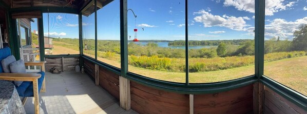 Ein Zimmer mit großem Fenster mit Blick auf einen See und Wald, Holzwände, einer Bank und einem an der Decke befestigten Vogelhaus.