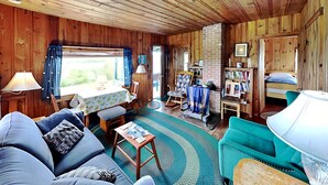A cozy cabin interior with a wooden ceiling, a dining area with a table and chairs, a sofa, a rocking chair, a bookshelf, and a bed.