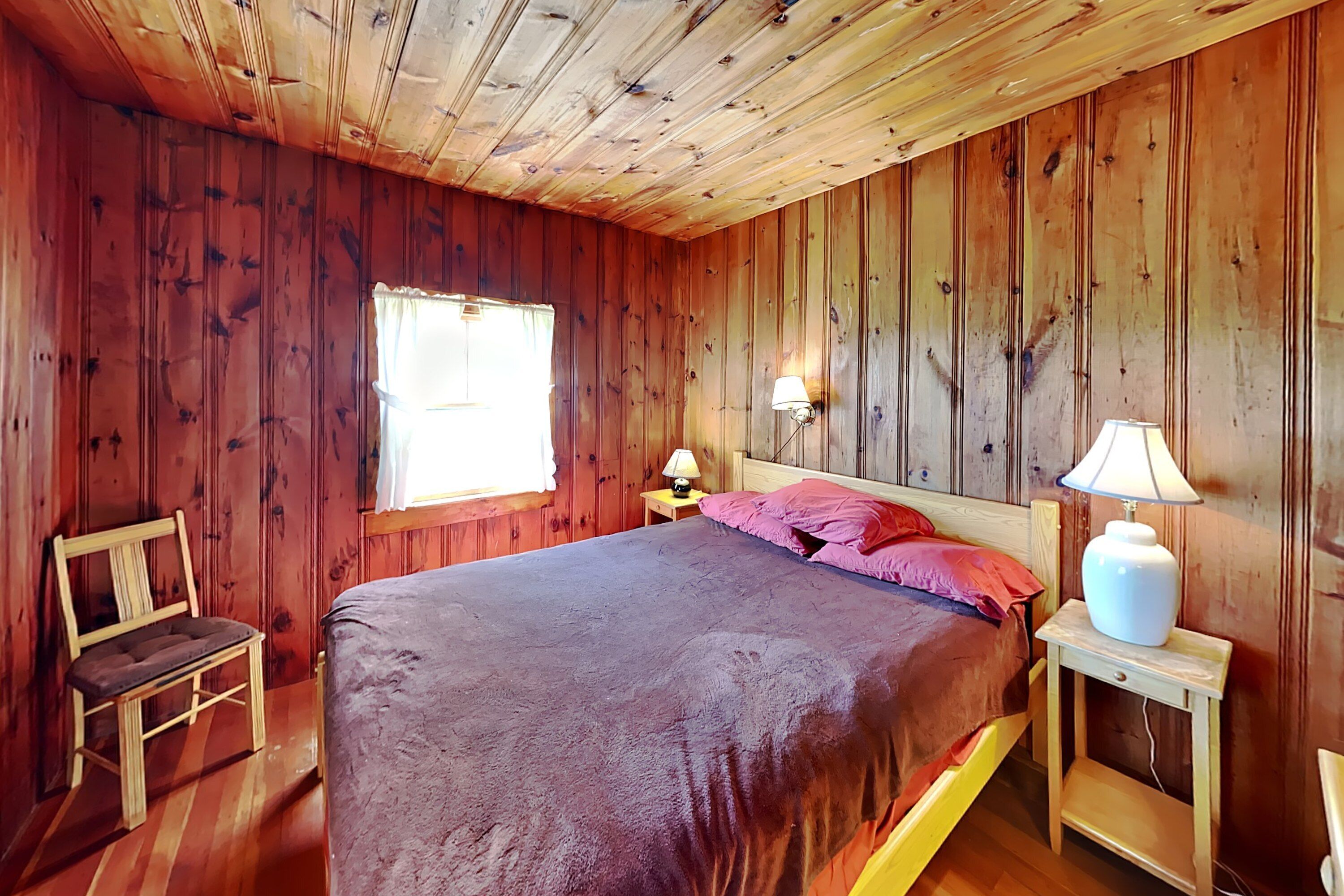 A wooden-paneled room with a bed, two bedside lamps, a chair, and a window.