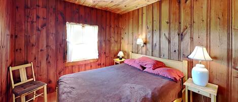 A wooden-paneled room with a bed, two bedside lamps, a chair, and a window.