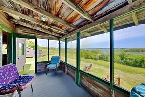 A covered porch with a view of a lake and greenery, wooden ceiling, and a chair with a patterned cushion. - Family-Friendly Castine Escape | Games, Kayaks & Views (Castine)