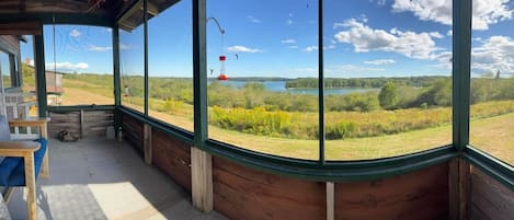 Ein Zimmer mit großem Fenster mit Blick auf einen See und Wald, Holzwände, einer Bank und einem an der Decke befestigten Vogelhaus.