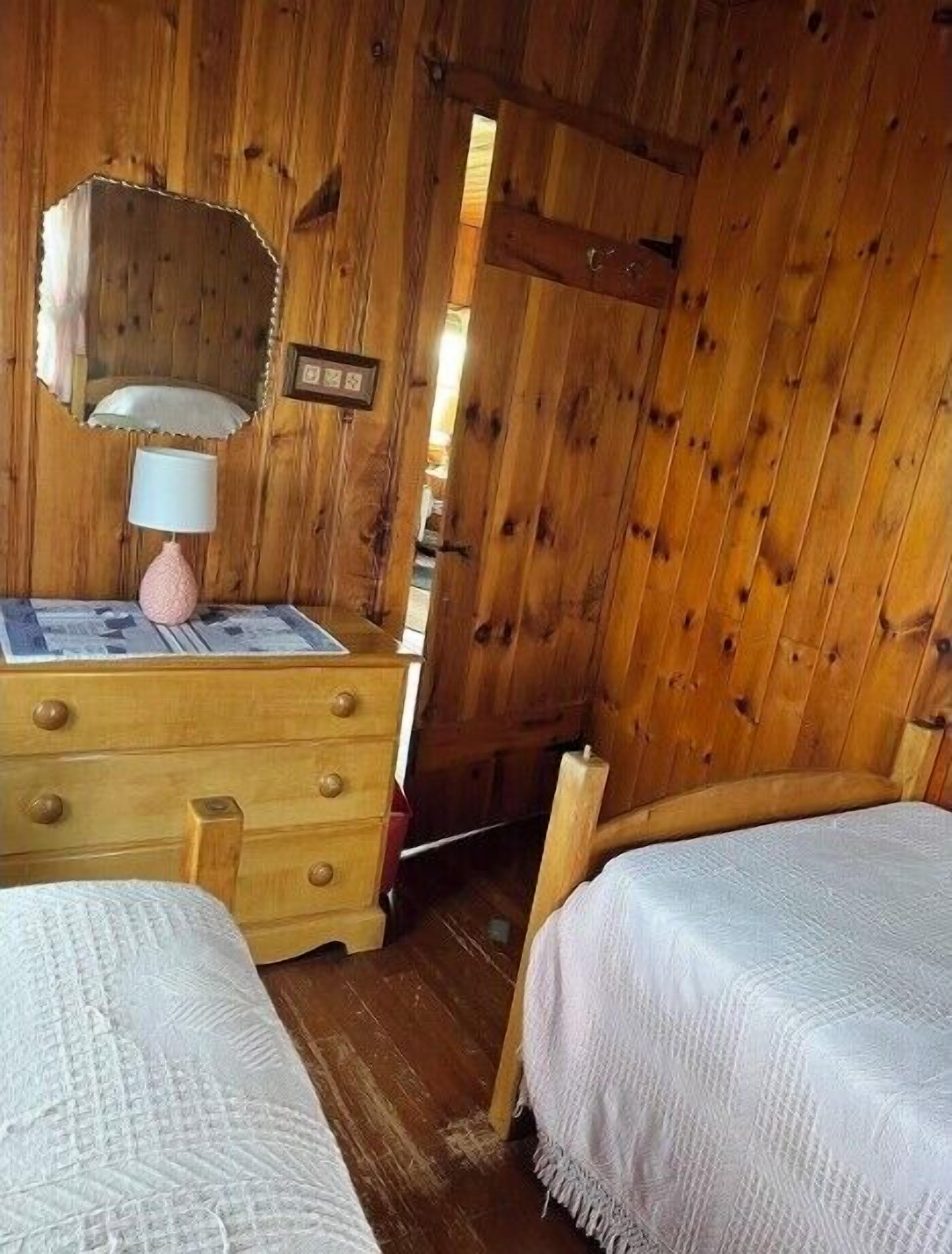 Quarto de cabana de madeira com cômoda, abajur, duas camas e espelho.