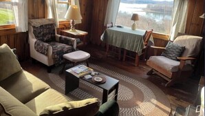 A cozy cabin interior with a sofa, armchair, dining table, and a view of a lake.