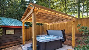 Un jacuzzi bajo una pérgola de madera con techo metálico, rodeado por una cerca de madera y un cobertizo pequeño.