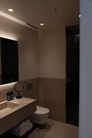 A modern bathroom with a beige countertop, a round mirror, a toilet, and a dark shower enclosure.