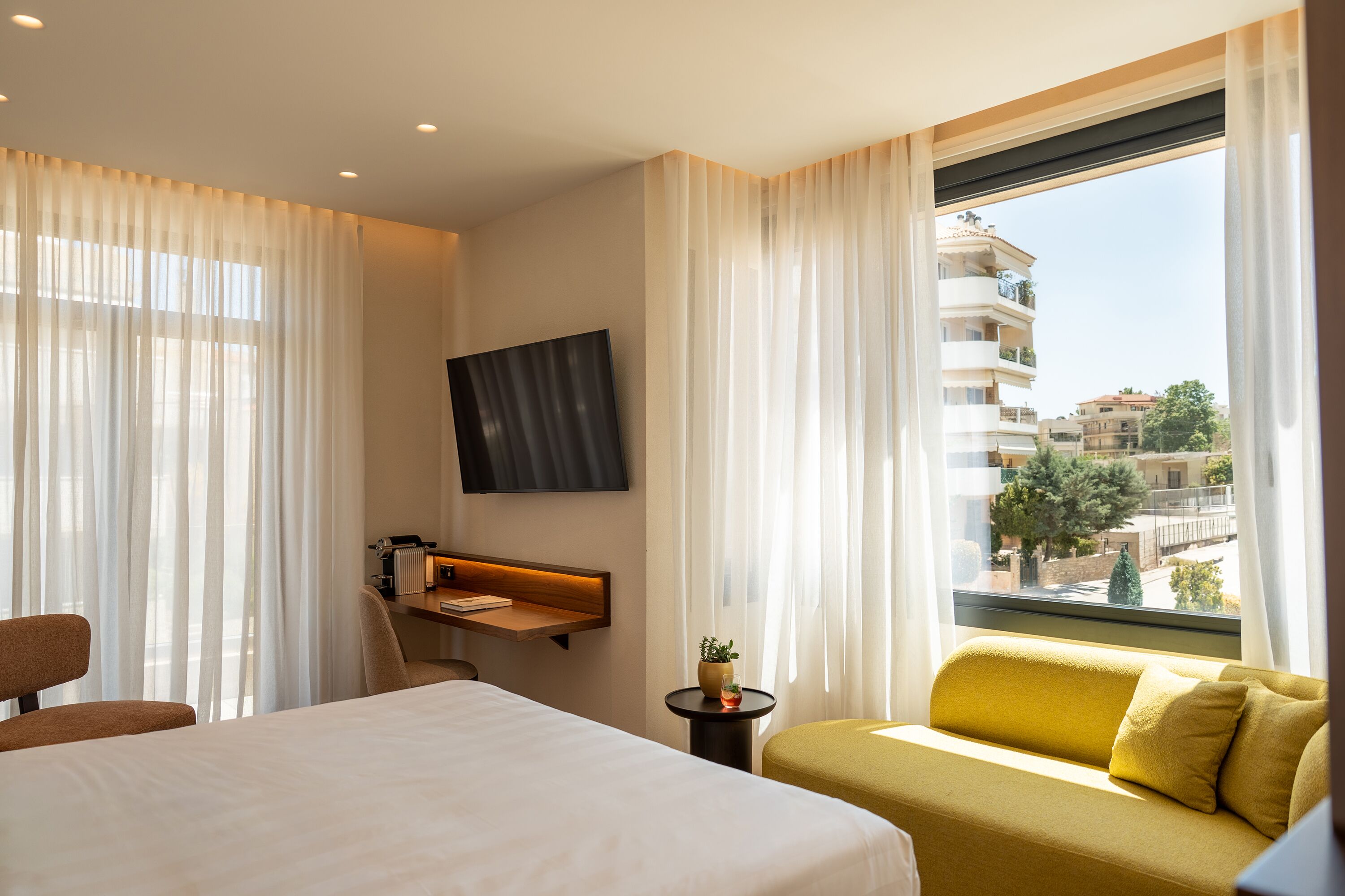 Habitación de hotel con cama, sofá, televisor en la pared, una mesita con planta y ventana con vistas a edificios.