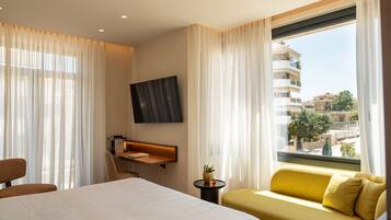 Habitación de hotel con cama, sofá, televisor en la pared, una mesita con planta y ventana con vistas a edificios.