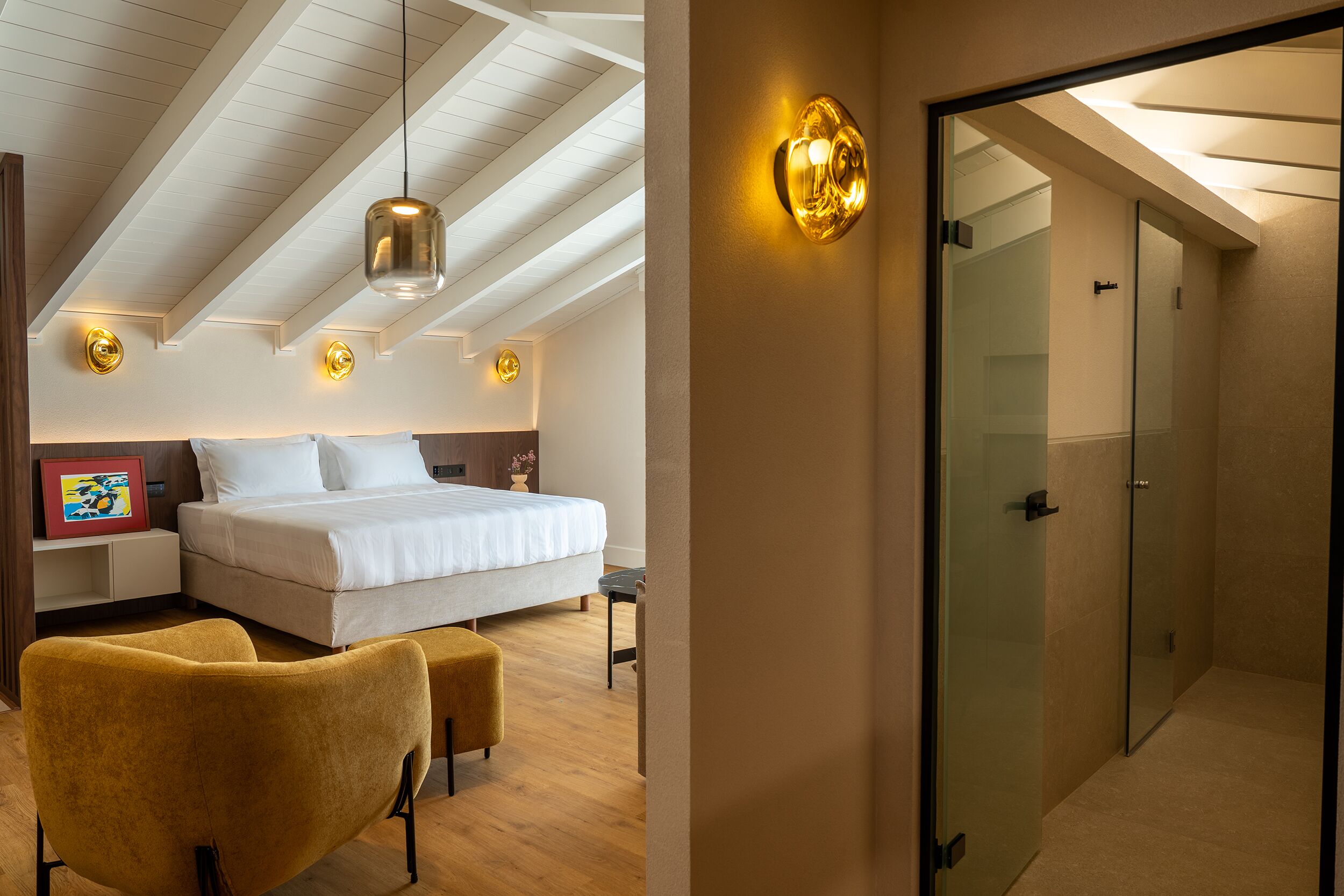 A bedroom with a bed, bedside tables, a chair, a hanging light fixture, and a glass door leading to a bathroom.