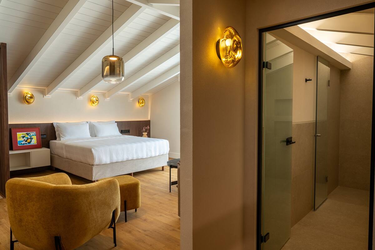 A bedroom with a bed, bedside tables, a chair, a hanging light fixture, and a glass door leading to a bathroom.