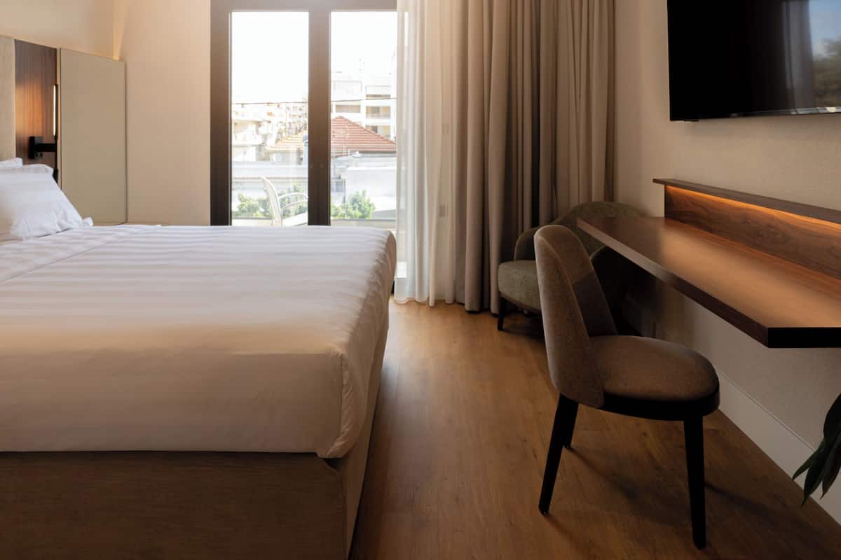 A hotel room with a bed, a desk, a chair, a flat-screen TV, and a large window with curtains.