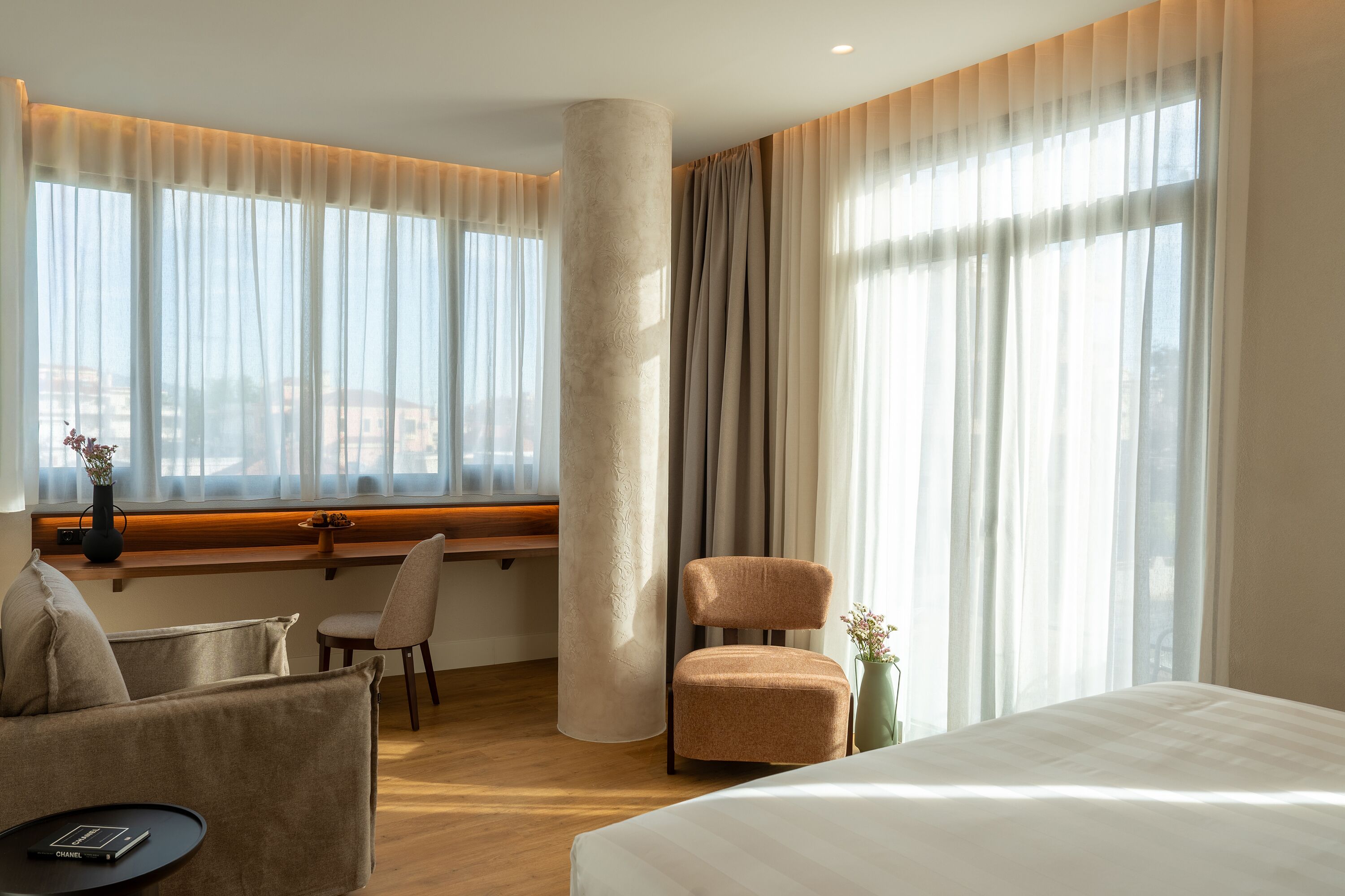 Habitación de hotel con cama, dos sillas, escritorio, ventana grande con cortinas translúcidas y vista a edificios.