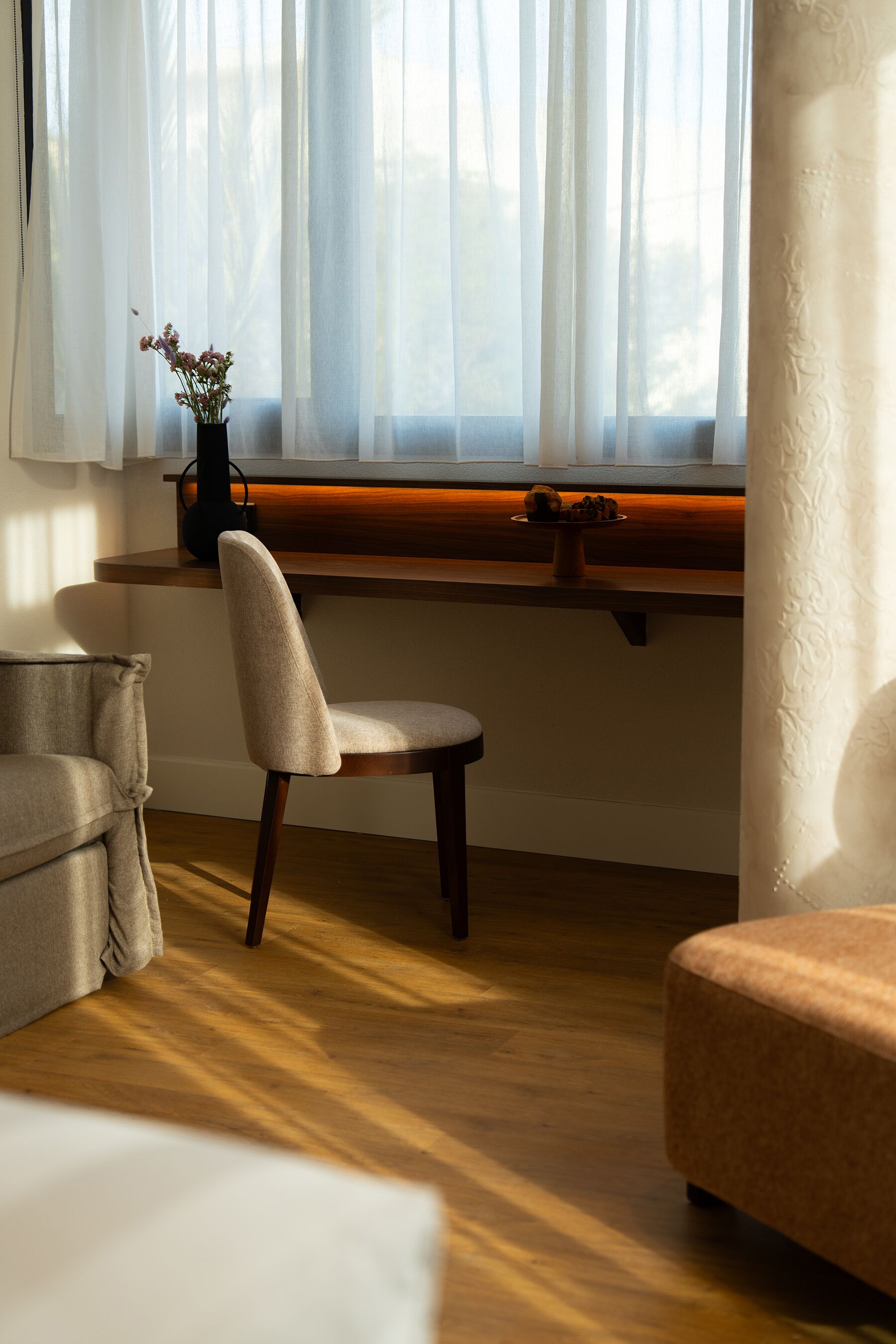 A room with a wooden floor, a window with sheer curtains, a chair, a sofa, and a small table with a vase of flowers.