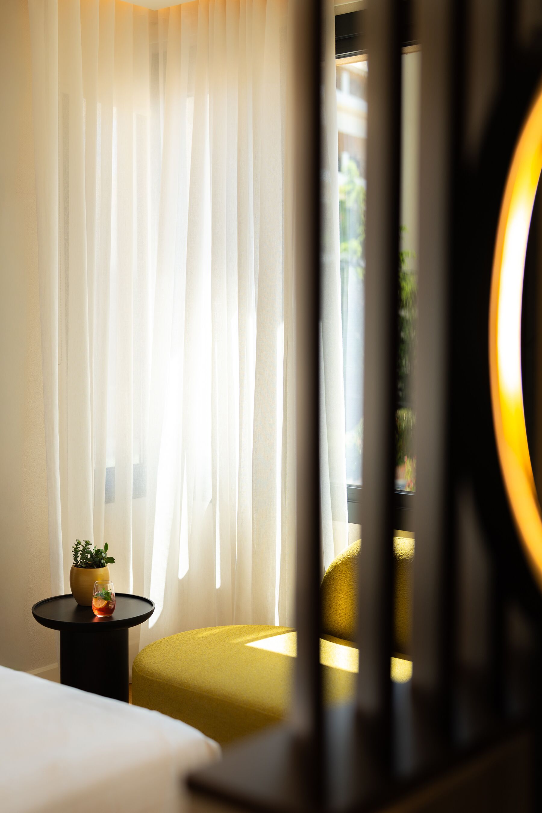 Een hotelkamer met een bed, een klein tafeltje met een plant, een geel voetenbankje en dunne gordijnen.