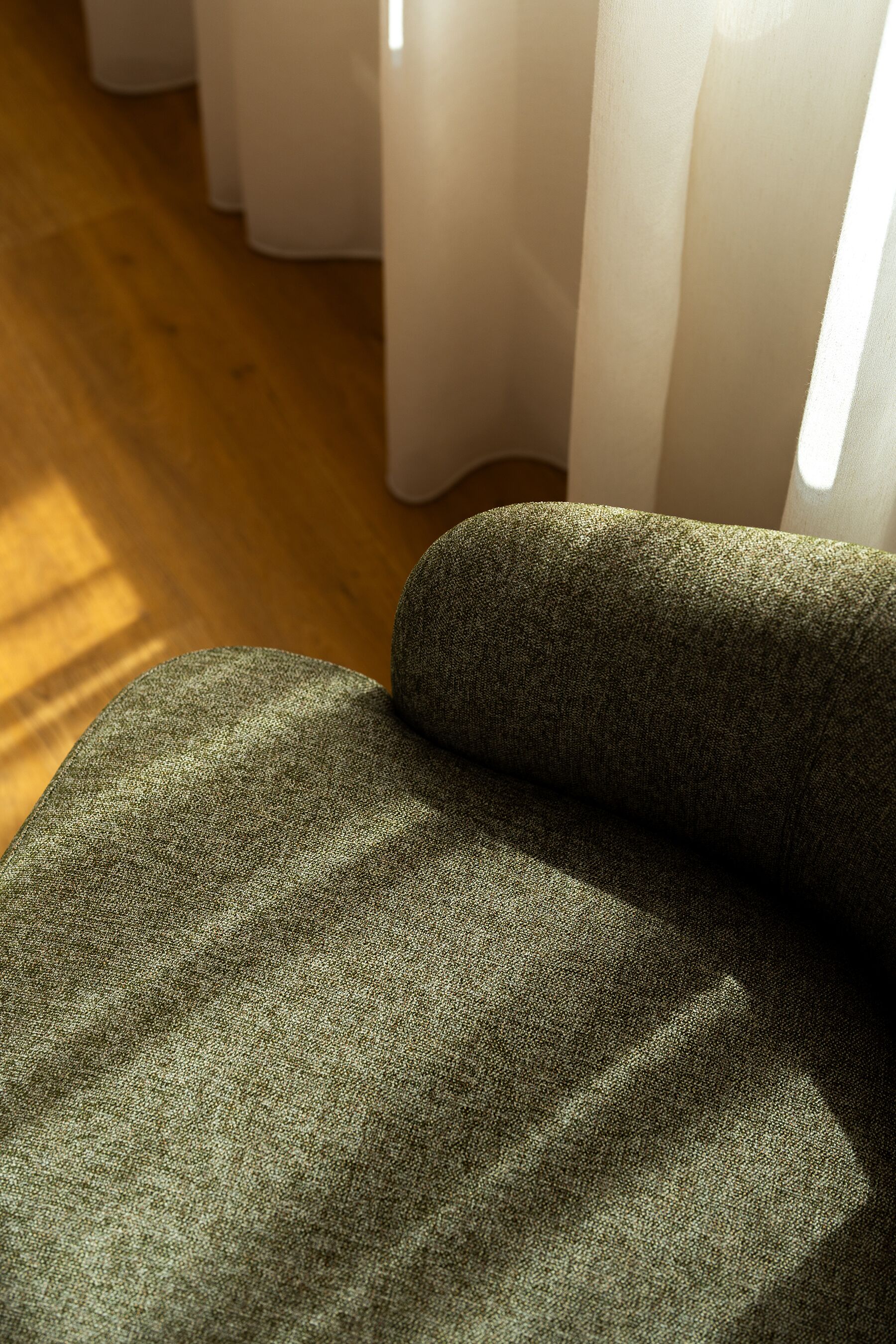 A close-up of a textured fabric, possibly a sofa or chair, with a wooden floor and white curtains in the background.