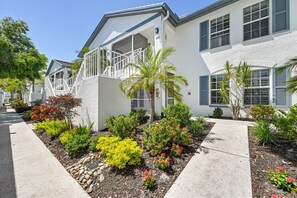 A modern two-story house with a balcony, a well-maintained garden with various plants, and a clear sky. - Renovated 2BR in Pelican Bay - Private Beach Access & Amenities (Naples)