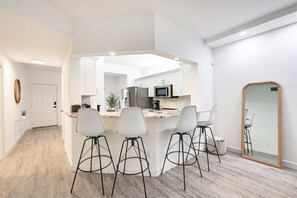 A modern kitchen with white cabinets, a stainless steel refrigerator, a microwave, and a large mirror. - Renovated 2BR in Pelican Bay - Private Beach Access & Amenities (Naples)