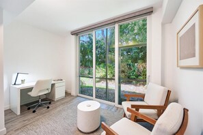 A modern room with a white desk, a chair, a round ottoman, and a large sliding glass door leading to a garden. - Renovated 2BR in Pelican Bay - Private Beach Access & Amenities (Naples)