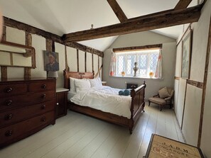 Ein Schlafzimmer mit einem hölzernen Bett, einem Schminktisch, einem Fenster mit Vorhängen und einem Sessel.
