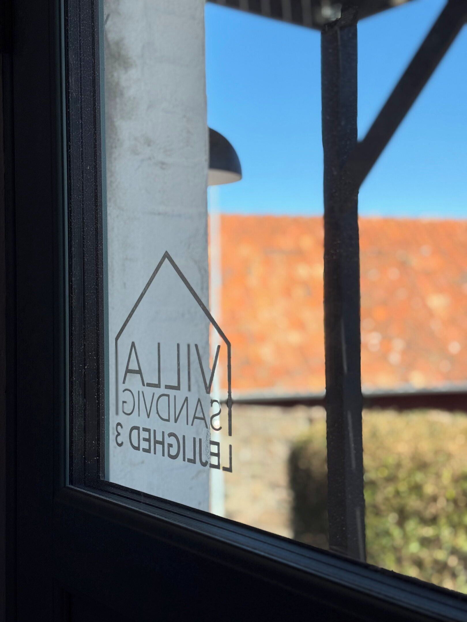 A window with a logo and text, a metal pole, and a blurred red-roofed building in the background.