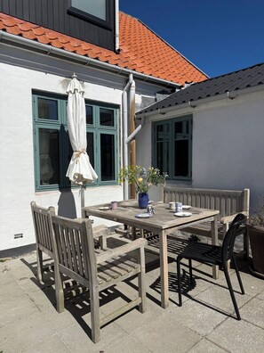 An outdoor patio with a wooden table and chairs, a potted plant, and a window with a curtain. - Holiday in Sandvig near sea and cliffs (Allinge-Sandvig)