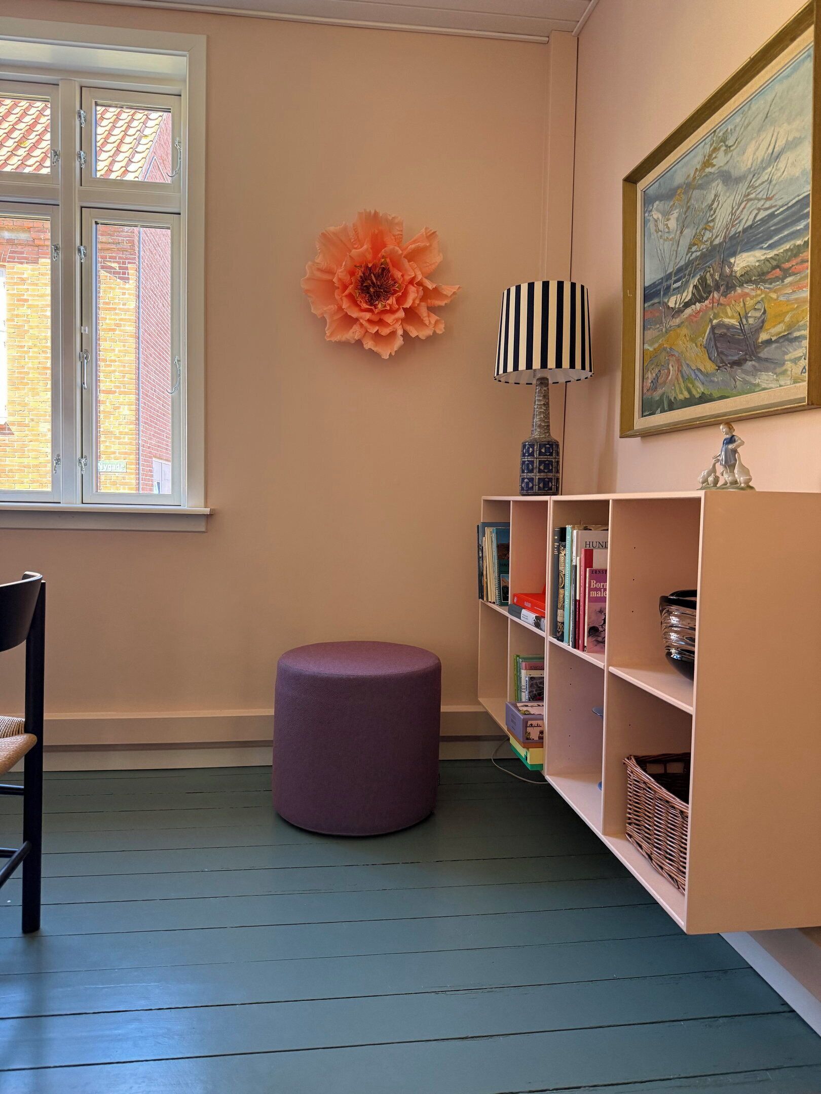 Una habitación con ventana, un reposapiés morado, una estantería y un cuadro en la pared.