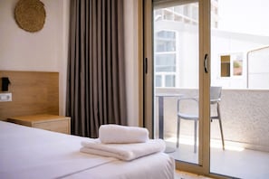 Habitación de hotel con cama, mesita de noche, luz en la pared, cortina y vistas al balcón exterior.