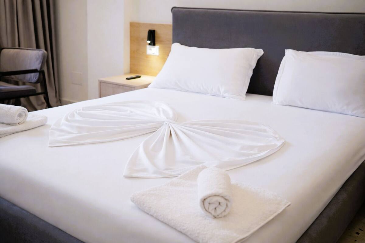 A neatly made bed with white linens, a remote control on the nightstand, and a chair in the background.
