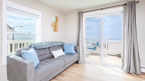 A living room with a grey sofa, a wooden floor, a large sliding glass door to a balcony, and a view of the sea. - Oceanfront Apartment w/ 2 Bedroom + den & pool access (Hampton)