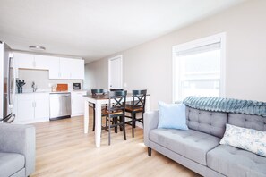 Un salon moderne avec un canapé gris, une table à manger avec des chaises et un coin cuisine doté de meubles blancs et d’appareils électroménagers.