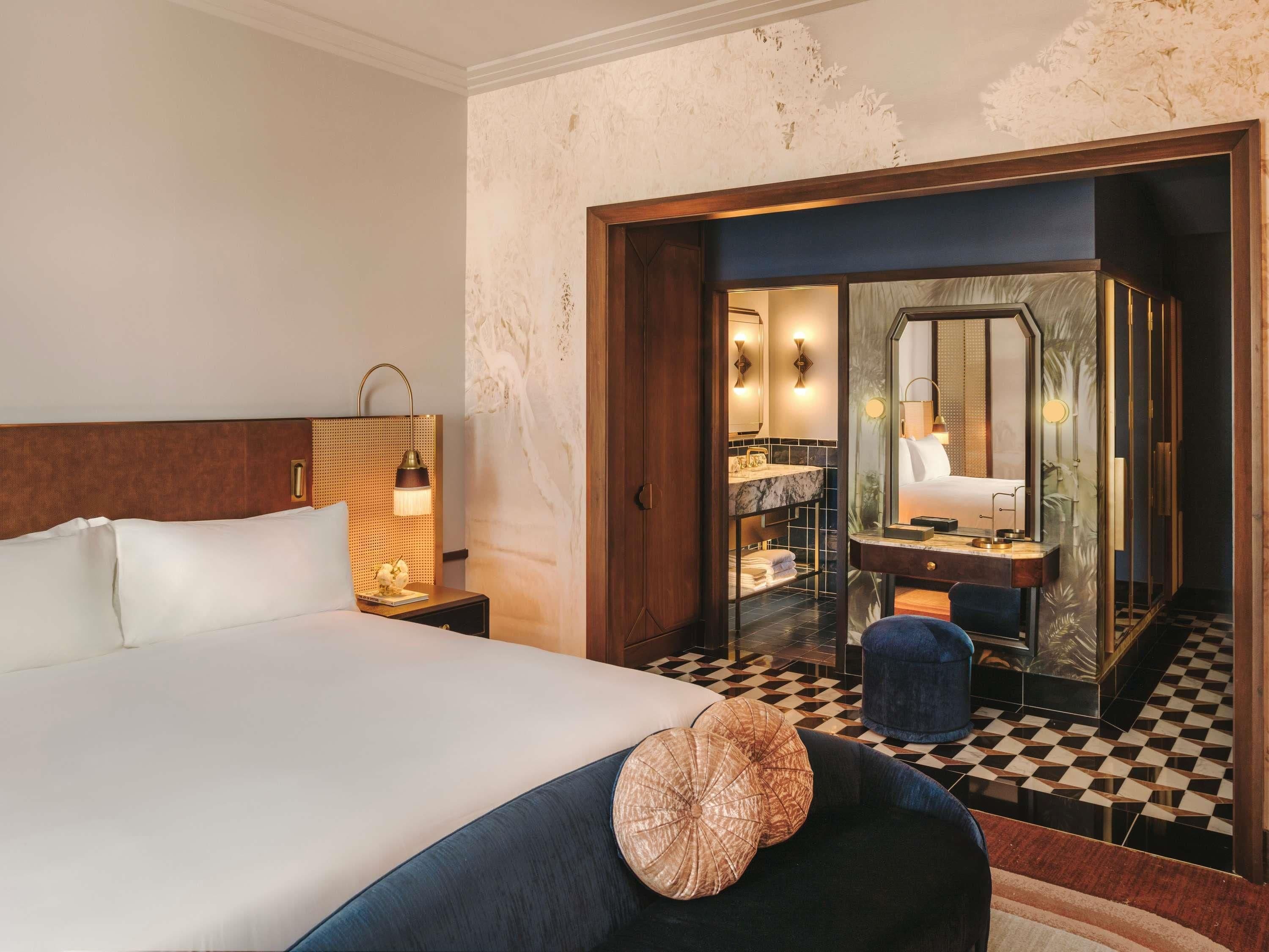 Une chambre d’hôtel avec un grand lit, une table de chevet avec une lampe, une salle de bain avec un miroir et un lavabo, et un sol à carreaux.