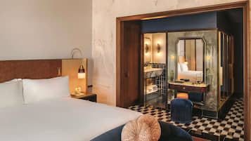 Une chambre d’hôtel avec un grand lit, une table de chevet avec une lampe, une salle de bain avec un miroir et un lavabo, et un sol à carreaux.