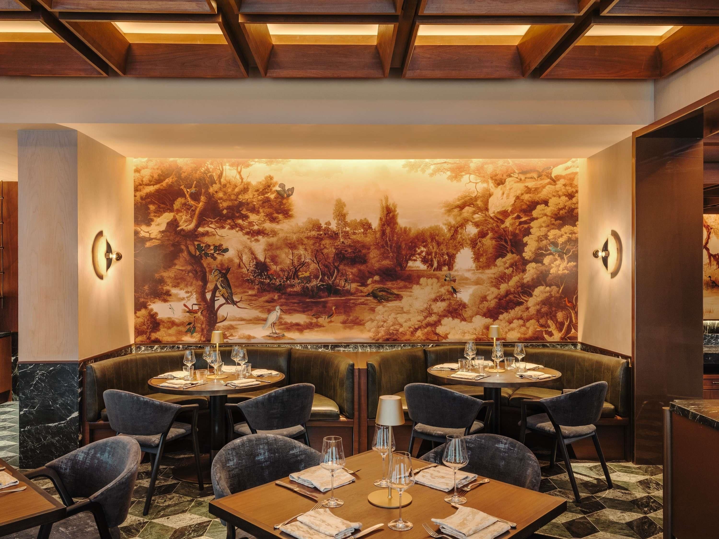 Un restaurant avec des tables rondes, des banquettes confortables et une grande fresque murale représentant une scène de forêt.