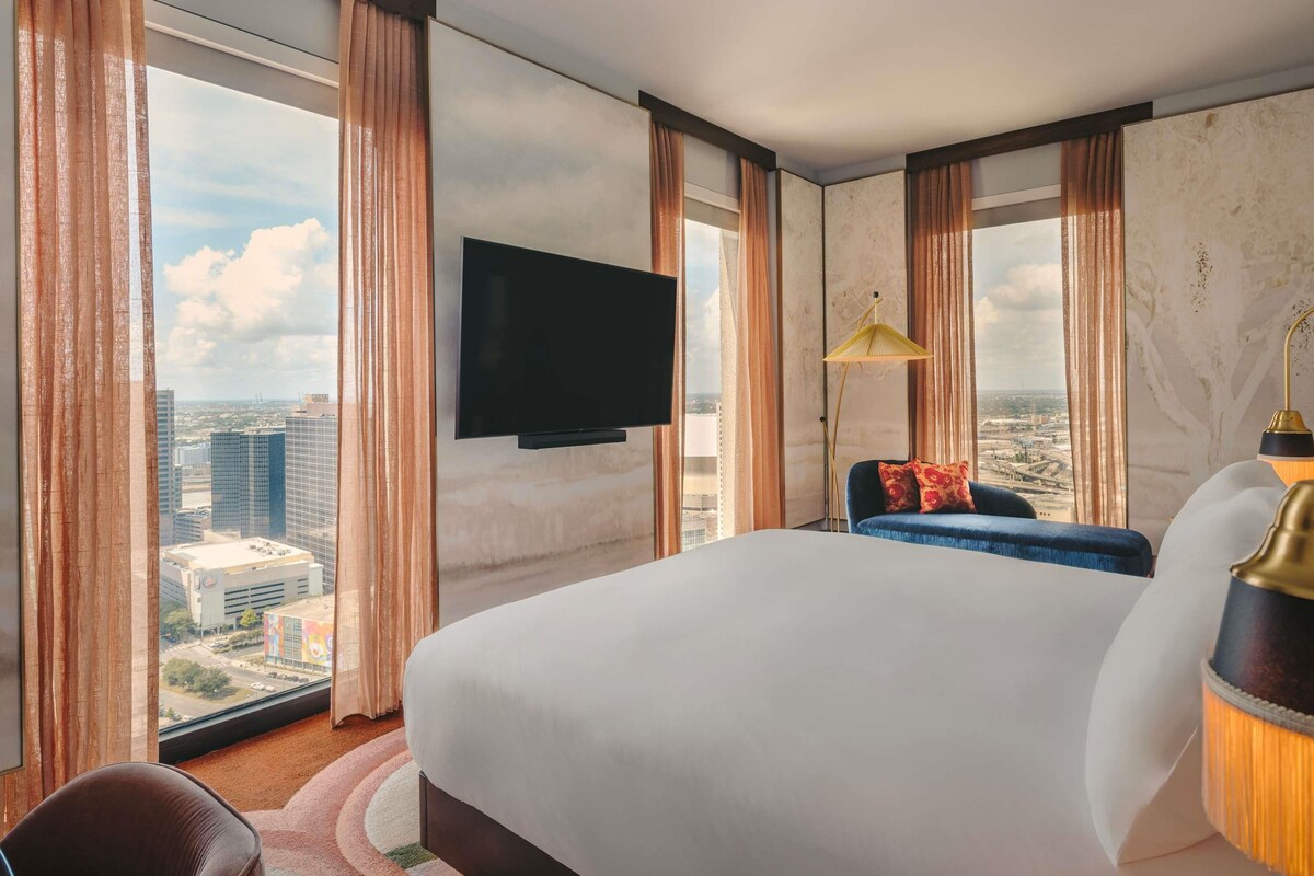 Habitación de hotel con una cama grande, un televisor de pantalla plana montado en la pared, un sofá con cojines, una lámpara de pie y vistas de la ciudad a través de amplios ventanales.